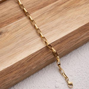 May include: A gold chain bracelet with a lobster clasp closure. The bracelet is made of a thick, boxy chain.