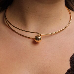 May include: A gold-colored choker necklace with a spherical pendant. The necklace features a double-strand design that curves around the neck, with a small, round ball as a focal point. The jewelry is simple and elegant.