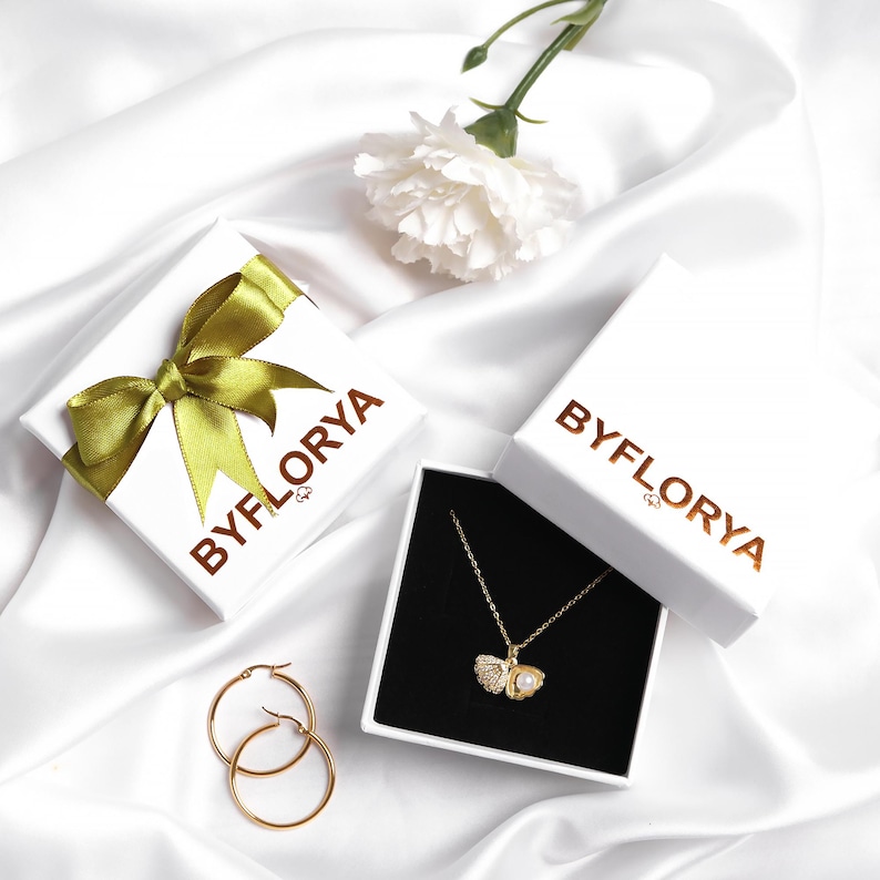 May include: A gold-toned necklace with a shell-shaped pendant containing a small white pearl, paired with two gold hoop earrings. The jewelry is presented in white boxes labeled BYFLORYA, one featuring a green ribbon, alongside a white flower.