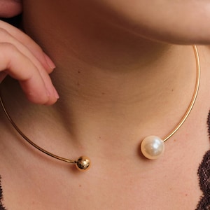 May include: A gold-coloured choker necklace with a large, round, white pearl at one end and a smaller, gold-coloured sphere at the other. The necklace is worn around the neck.