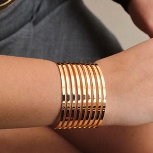 May include: A collection of gold-colored bangles stacked on a wrist. The bracelets are smooth and shiny, reflecting light. The image shows a close-up of the bracelets, highlighting their metallic sheen and simple design. The bangles are a classic jewelry item.