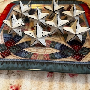 May include: A collection of eight white star-shaped cabinet knobs with black distressed edges. The knobs are arranged on a decorative tray with a blue and red border and a floral pattern. The knobs are made of metal.