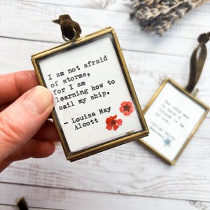 Può includere: Un ornamento rettangolare con cornice dorata con la citazione "I am not afraid of storms, for I am learning how to sail my ship" di Louisa May Alcott. Lo sfondo bianco presenta accenti floreali rossi. Un secondo ornamento è visibile.