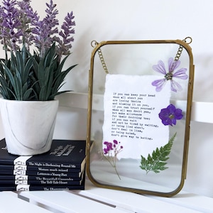 May include: A rectangular brass-framed glass display featuring a piece of white paper with black printed text from the poem If by Rudyard Kipling, decorated with pressed purple flowers, a small green fern leaf, and tiny pink blossoms.