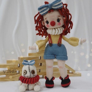 Puede incluir: Una muñeca de crochet con pelo rojo, un lazo azul y blanco a rayas y una camisa amarilla con un mono azul. La muñeca sostiene una muñeca de crochet más pequeña con un cuello rojo y un lazo azul. Las muñecas están de pie sobre una superficie de madera.