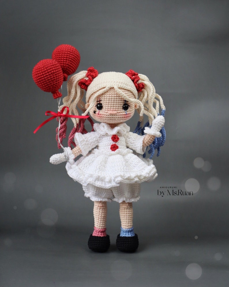 Puede incluir: Una mu&ntilde;eca de crochet con cabello rubio, vestida con un vestido blanco con detalles rojos y sosteniendo dos globos rojos. La mu&ntilde;eca tiene una l&aacute;grima dibujada en la mejilla y lleva zapatos negros. La mu&ntilde;eca est&aacute; etiquetada como "Amigurumi by MsRutan".
