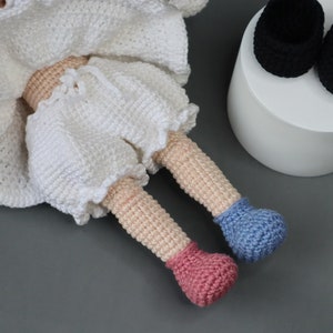 Puede incluir: Una mu&ntilde;eca de crochet con ropa blanca y zapatos de crochet rosa y azul. La mu&ntilde;eca est&aacute; sentada sobre una superficie gris.