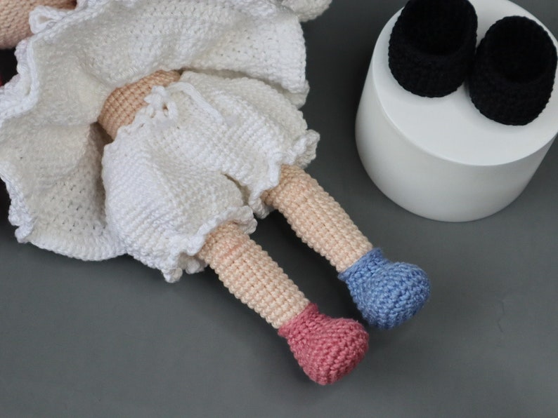 Puede incluir: Una mu&ntilde;eca de crochet con ropa blanca y zapatos de crochet rosa y azul. La mu&ntilde;eca est&aacute; sentada sobre una superficie gris.