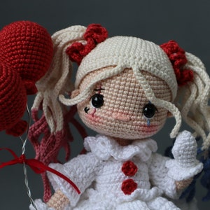 Puede incluir: Una mu&ntilde;eca de crochet con cabello blanco, un vestido blanco con detalles rojos y una l&aacute;grima en la mejilla. Est&aacute; sosteniendo dos globos rojos.