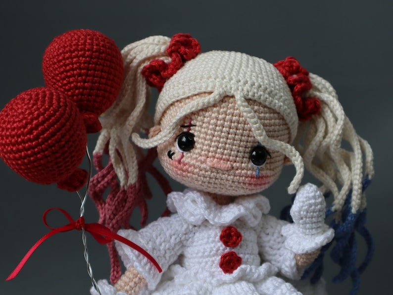Puede incluir: Una mu&ntilde;eca de crochet con cabello blanco, un vestido blanco con detalles rojos y una l&aacute;grima en la mejilla. Est&aacute; sosteniendo dos globos rojos.