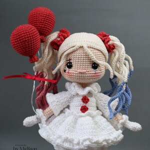Puede incluir: Una mu&ntilde;eca de crochet con el pelo rubio en trenzas, que lleva un vestido blanco con botones rojos y sostiene dos globos rojos. La mu&ntilde;eca tiene una l&aacute;grima dibujada en la mejilla y lleva guantes blancos.