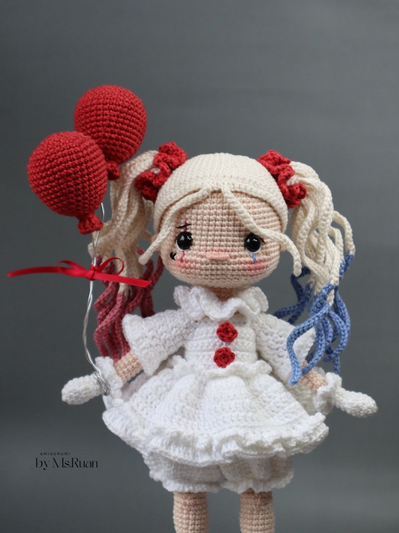 Puede incluir: Una mu&ntilde;eca de crochet con el pelo rubio en trenzas, que lleva un vestido blanco con botones rojos y sostiene dos globos rojos. La mu&ntilde;eca tiene una l&aacute;grima dibujada en la mejilla y lleva guantes blancos.