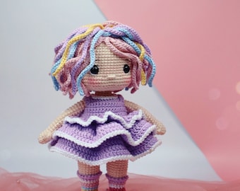 Pode incluir: Uma boneca de crochê com um vestido roxo, sapatos rosa e cabelo de lã arco-íris.