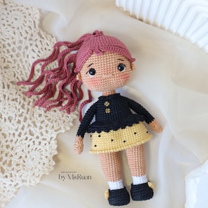 May include: A crocheted doll with pink hair, a black sweater with gold buttons, a yellow skirt with black polka dots, and black shoes. The doll is sitting on a white surface with a crocheted doily.