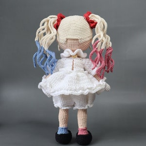 Puede incluir: Una mu&ntilde;eca de crochet con cabello rubio, detalles rojos y azules y un vestido blanco. La mu&ntilde;eca lleva zapatos negros y tiene un cuello de crochet blanco.