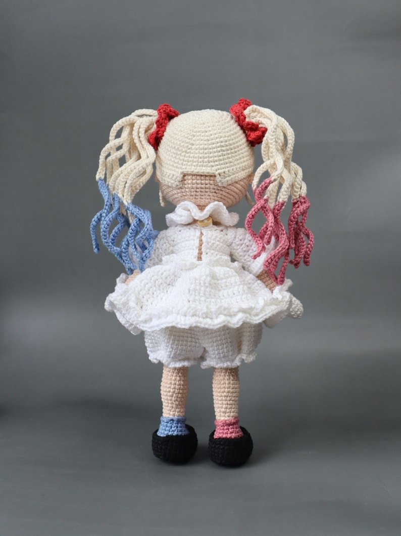 Puede incluir: Una mu&ntilde;eca de crochet con cabello rubio, detalles rojos y azules y un vestido blanco. La mu&ntilde;eca lleva zapatos negros y tiene un cuello de crochet blanco.