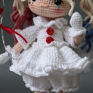 Puede incluir: Una mu&ntilde;eca de crochet que lleva un vestido blanco con botones rojos y un volante blanco. La mu&ntilde;eca tiene el pelo rubio y los ojos azules. La mu&ntilde;eca sostiene un globo rojo.