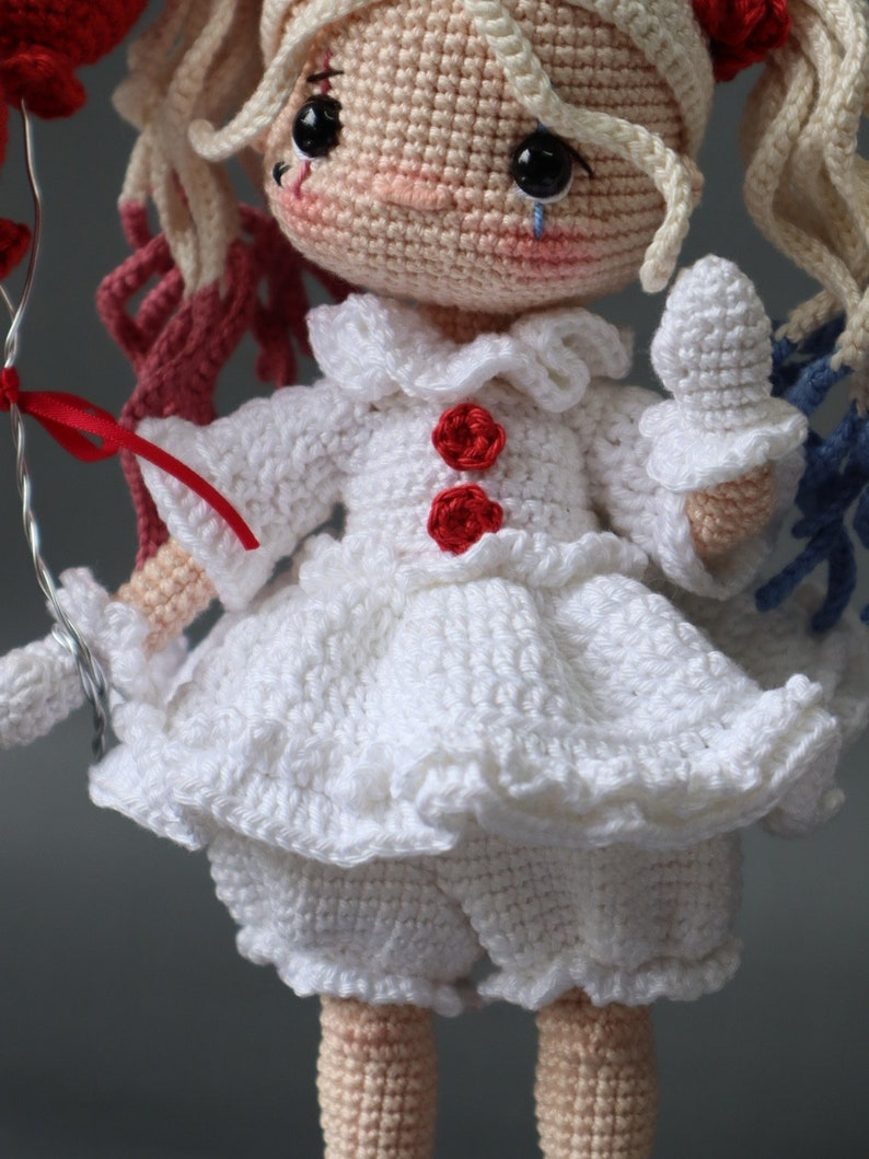 Puede incluir: Una mu&ntilde;eca de crochet que lleva un vestido blanco con botones rojos y un volante blanco. La mu&ntilde;eca tiene el pelo rubio y los ojos azules. La mu&ntilde;eca sostiene un globo rojo.