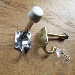 May include: Two door stops, one chrome and one brass, with rubber stoppers. Both have a hook on the bottom. The chrome door stop has a white rubber bumper. The brass door stop has a black rubber bumper. The door stops are mounted on a wooden surface.