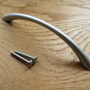 Może przedstawiać: Srebrny uchwyt do drzwi szafki o zakrzywionym kształcie. W zestawie dwa wkręty do montażu.