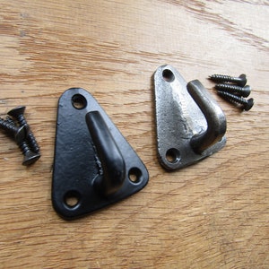 Puede incluir: Dos ganchos de metal triangulares con un gancho curvo en el extremo. Un gancho es negro y el otro es plateado. Ambos ganchos tienen dos agujeros para tornillos. Los ganchos están sobre una superficie de madera.
