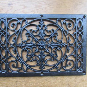 Può includere: Copertura di ventilazione decorativa in ghisa nera con un design a volute ornato e simmetrico. Il coperchio rettangolare ha un centro floreale ed è mostrato con viti su una superficie di legno.