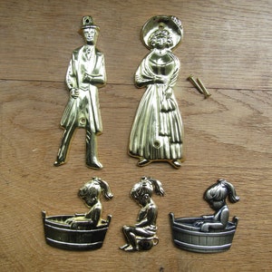 May include: A set of five gold-toned metal figurines depicting a couple and two children. The couple is dressed in Victorian-era clothing, and the children are in bathing suits. The figurines are attached to a wooden surface.