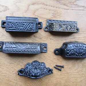 May include: Five antique drawer pulls with ornate designs. Four are black metal with intricate floral patterns. One is gold metal with a stylized floral design. Two screws are included.