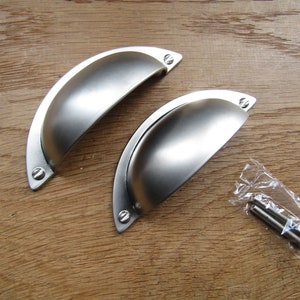 以下が含まれることがあります： 丸みを帯びた、わずかに湾曲した形状の銀色のキャビネットノブが2つ。ノブはブラッシュ仕上げで、木製の表面に取り付けられています。透明なプラスチック袋に2本のネジが付属しています。