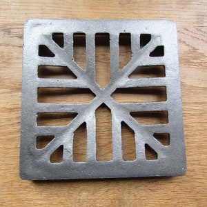 Könnte beinhalten: Ein quadratischer, gusseiserner Abwasserkanalrost mit einem dekorativen, geometrischen Design. Der Rost ist silberfarben und hat eine leicht strukturierte Oberfläche.