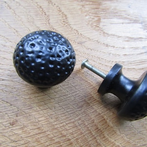 May include: Two black cabinet knobs with a textured, hammered finish. One knob is a round ball and the other is a round ball with a stem.