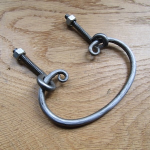 Op de afbeelding: Een zilveren metalen ladegreep met een gedraaid ontwerp en twee schroeven. De greep bevindt zich op een houten oppervlak.