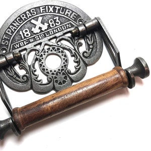 Könnte beinhalten: Ein Toilettenpapierhalter aus Gusseisen mit einer Holzrolle. Der Halter hat ein verziertes Design und den Text "St Pancras Fixture 1883 WBF & SIO LONDON".