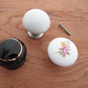 Può includere: Tre pomelli per armadi in ceramica. Uno è bianco, uno è nero con un bordo dorato e uno è bianco con un motivo floreale.