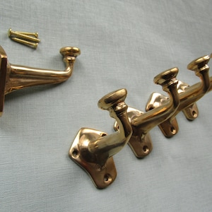May include: Set of brass wall hooks with a decorative design. The hooks have a curved arm and a rounded end. Includes mounting screws. The hooks are approximately 4 inches long.