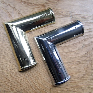 Puede incluir: Dos protectores de esquina, uno en latón pulido y otro en cromo, sobre una superficie de madera.