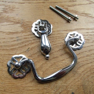 Puede incluir: Dos tiradores de armario de níquel pulido con diseños florales ornamentados. El tirador más grande es una manija de caída con una forma curva. El tirador más pequeño es un pomo redondo.