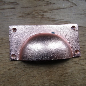 May include: A copper-coloured cabinet pull with a rounded, recessed centre. The pull has four screw holes.