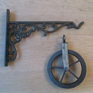 Peut inclure: Un support mural en métal gris foncé présentant un motif à volutes ornementales avec un crochet à l'extrémité, accompagné d'une poulie circulaire séparée avec une pièce de support centrale en métal et une petite boucle sur le dessus.