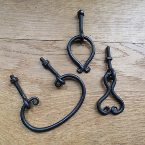May include: Three black metal drawer pulls with a unique, hand-forged design. Each pull has a different shape, with one featuring a heart shape, another a rounded loop, and the third a teardrop shape.