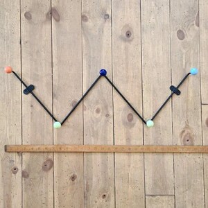 Könnte beinhalten: Ein schwarzer Metall-Kleiderständer mit fünf Haken. Die Haken sind mit farbigen Kugeln verziert: orange, blau, grün und zwei hellblau. Der Kleiderständer ist an einer Holzwand montiert.