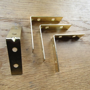 May include: Four gold metal corner brackets with four holes each. The brackets are arranged in a staggered pattern on a wooden surface.