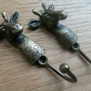 May include: Two antique gold giraffe-shaped hooks with a spotted pattern. The hooks are attached to a wooden surface.