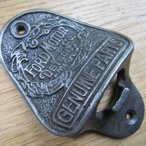 Op de afbeelding: Een flessenopener van gietijzer met de tekst "Ford Motor Co. Detroit, Mich. Genuine Parts" in reliëf. De opener is zilverkleurig en heeft een versleten, vintage uitstraling.