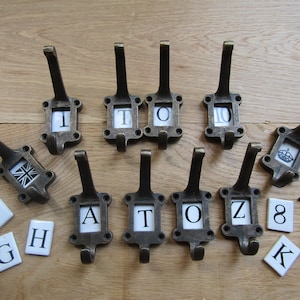 May include: A set of ten antique-style hooks with a variety of letter and number tiles. The hooks are made of metal and have a dark brown finish. The tiles are white with black lettering. The tiles include the letters A, T, O, Z, G, H, K, and the numbers 1, 8, and 10.