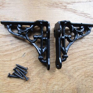 May include: Two black metal shelf brackets with a decorative scroll design. The brackets are shown on a wooden surface with screws.
