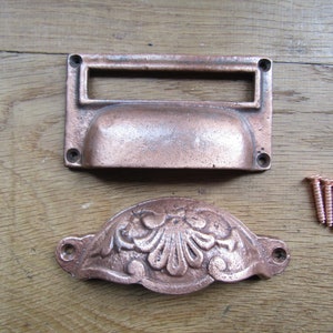 Puede incluir: Dos tiradores de cajón de cobre antiguo con un diseño decorativo. Un tirador tiene una ranura rectangular para una etiqueta. Los tiradores se acompañan de tres tornillos.