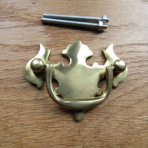 May include: A brass cabinet pull with a decorative, ornate design. The pull has a loop handle and two decorative knobs on either side. The pull is shown on a wooden surface with two screws.