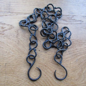 Könnte beinhalten: Eine schwarze Metallkette mit dekorativen Gliedern und Haken an jedem Ende. Die Kette liegt auf einer Holzoberfläche.