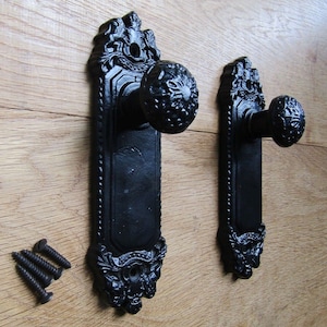 May include: Two black cast iron door knobs with ornate detailing. The knobs have a traditional design with a round handle and a decorative plate. The knobs are mounted on a wooden surface.